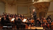 Auf der Bühne der Hamburger Laeiszhalle steht ein Dirigent bei einem Konzert vor einem Orchester. © Screenshot 