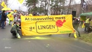 Teilnehmende einer Protestaktion halten ein gelbes Banner mit der Aufschrift: "Atomausstieg muss geLingen". Eine Person streut ein gelbes Pulver auf die Straße. © Screenshot 