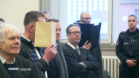 Im Gerichtsaal: Angeklagte halten sich Mappen vors Gesicht, um nicht erkannt zu werden. © Screenshot 