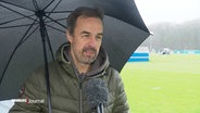 Reporter Lars Pegelow neben dem Fußballplatz unterm Regenschirm. © Screenshot 