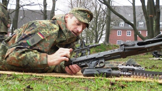 Logistiker Frank liegt in Tarnkleidung auf dem Bauch auf einer Wiese und lädt ein Gewehr. © Screenshot 