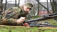 Logistiker Frank liegt in Tarnkleidung auf dem Bauch auf einer Wiese und lädt ein Gewehr. © Screenshot 