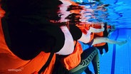 Unterwasseraufnahme von Kindern mit orangenen Schwimmwesten in einem Hallenbad. © Screenshot 