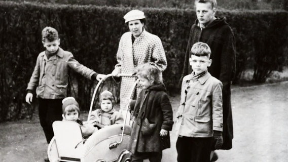 Eine alte schwarz-weiß-Aufnahme: Eine Frau schiebt einen Kinderwagen mit zwei Kleinkindern. Sie ist umgeben von drei weiteren größeren Kindern. © Screenshot 
