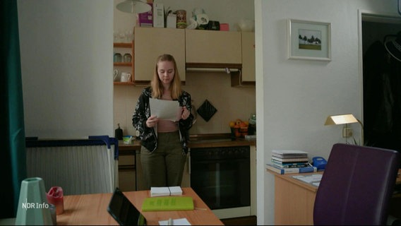 Eine junge Frau steht in einer kleinen Wohnung und liest ein Schreiben, das sie in der Hand hält. © Screenshot 