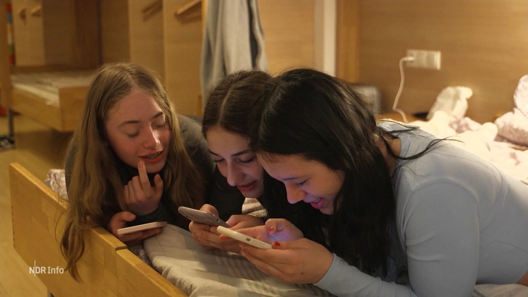 Drei Mädchen liegen nebeneinander im Bett und schauen auf ihre Handys.