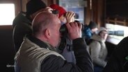 Menschen mit Ferngläsern und Kameras in einer Hütte mit Fensterluken. © Screenshot 