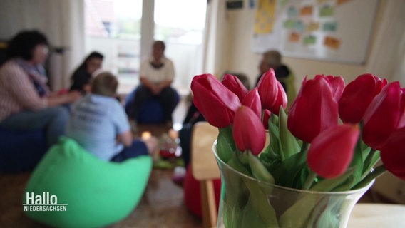 Tulpen in einer Vase, im Hintergrund eine sitzende Menschengruppe. © Screenshot 