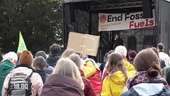 Demonstrierende vor einer Bühne, auf der eine Kundgebung stattfindet. Auf einem Transparent an der Bühne steht "End Fossil Fuels Now". © Screenshot 