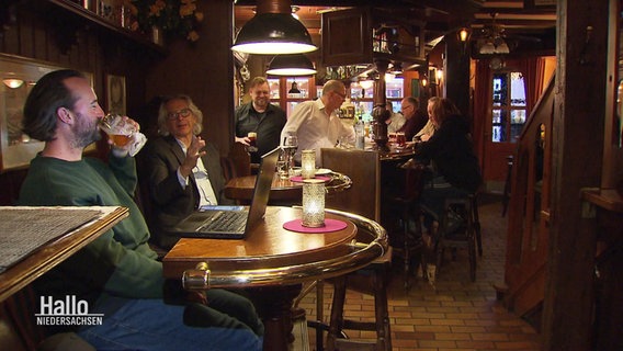 Szene in einer Kneipe: Im Vordergrund sitzen zwei Männer an einem Tisch mit einem Laptop und trinken Bier, im Hintergrund werden mehrere Personen am Tresen bedient. © Screenshot 