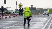 Polizisten haben eine Straße abgesperrt und halten in neongelben Jacken und mit Polizeikellen Autos an. © Screenshot 