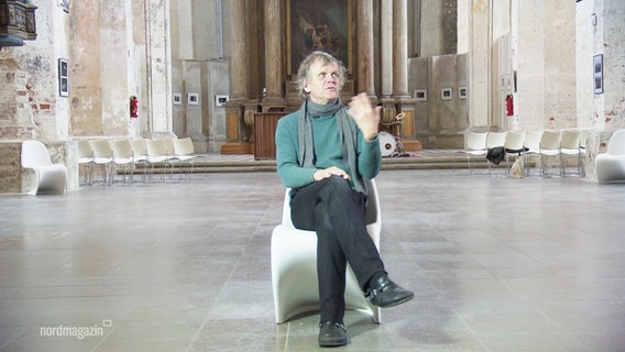 Moritz Götze, Organisator der Ausstellung, sitzt auf einem weißen Stuhl. © Screenshot 