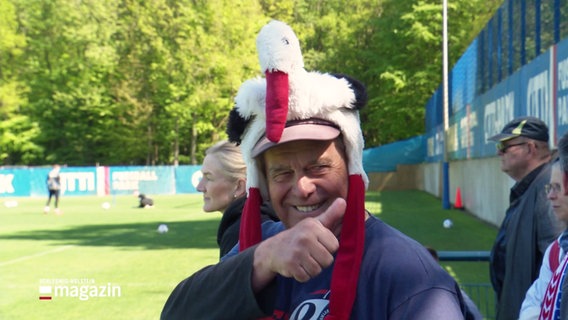 Ein Fan von Holstein Kiel mit einer Mütze in Form eines Storchs am Spielfeldrand beim Training. Er reckt einen Daumen in die Höhe und grinst. © Screenshot 