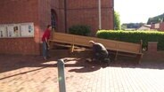 Zwei Männer stellen eine lange Kirchenbank vor der Kirche ab. © Screenshot 