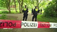 Polizisten stehen in einem Park hinter rot-weißem Absperrband. © Screenshot 