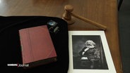 Die Erstausgabe von "Das Kapital" von Karl Marx, daneben eine Lupe, ein Foto des Autors sowie der Hammer einer Auktion liegen auf einem Schreibtisch. © Screenshot 