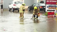 Starkregen: Zwei Mitarbeiter der Feuerwehr stehen auf einer Straße fast knietief im Wasser. © Screenshot 