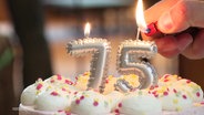 Nahaufnahme einer Geburtstagstorte. Darauf zwei Kerzen in der Form einer 75. Die Flamme auf der 7 brennt schon, die auf der 5 wird gerade von einer Hand mit Feuerzeug angezündet. © Screenshot 