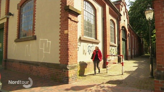 Eine Frau in roter Jacke läuft an einem alten Backsteingebäude entlang. © Screenshot 