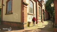 Eine Frau in roter Jacke läuft an einem alten Backsteingebäude entlang. © Screenshot 