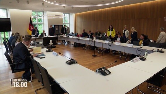 Ein Plenarsaal mit einem Tischkreis und vielen Menschen, die stehen. © Screenshot 