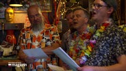 Männer in Hawaii-Hemden und künstlichen Blumenketten stehen dicht beieinander und singen. In ihren Händen halten sie Papier, auf dem offenbar der Liedtext steht. © Screenshot 