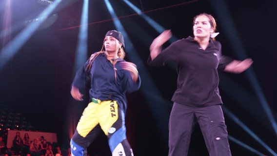 Zwei Frauen stehen auf einer Bühne und tanzen, im Hintergrund sitzt Publikum. © Screenshot 