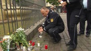 Blumensträuße und Kerzen liegen auf dem Boden vor einem Zaun. Zwei Polizeibeamte hocken bzw. stehen davor und entzünden weitere Kerzen. © Screenshot 