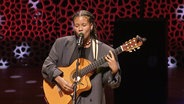 Die Musikerin Ami Warning auf der Bühne der Elbphilharmonie. Sie ist eine junge Frau mit dunklen, geflochteten Zöpfen. Sie singt in das Mikrofon und spielt dazu selbst auf einer Gitarre. © Screenshot 