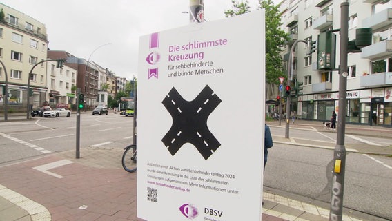 Vor einer großen Kreuzung hängt ein Plakat mit der Aufschrift "Schlimmste Kreuzung für sehbehinderte und blinde Menschen". © Screenshot 