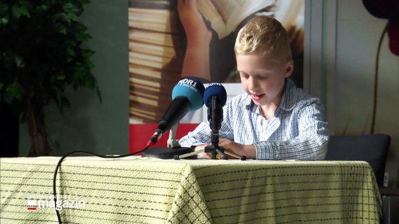 Ein Junge im Grundschulalter sitzt an einem Tisch vor einem Mikrofon und liest etwas vor. © Screenshot 