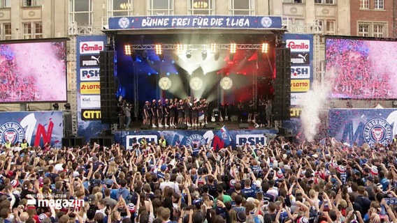 Fans von Holstein Kiel jubeln vor einer Bühne während einer Aufstiegsfeier. © Screenshot 