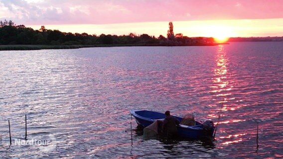 Bei Sonnenaufgang schwimmt ein kleines Beiboot in einer Bucht neben einem Krabbenfischer in Wathose. © Screenshot 