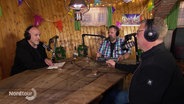 Drei Männer sitzen in einem Partykeller an einem Tisch vor Mikrophonen und unterhalten sich. © Screenshot 