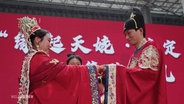 Ein junges Paar in roten, reich verzierten Gewändern reicht sich bei einer traditionellen chinesischen Hochzeit die Hände. © Screenshot 