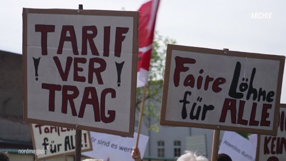 Plakate mit der Aufschrift "Tarifvertrag" und "Faire Löhne für Alle" werden auf einer Demo hochgehalten. © Screenshot 