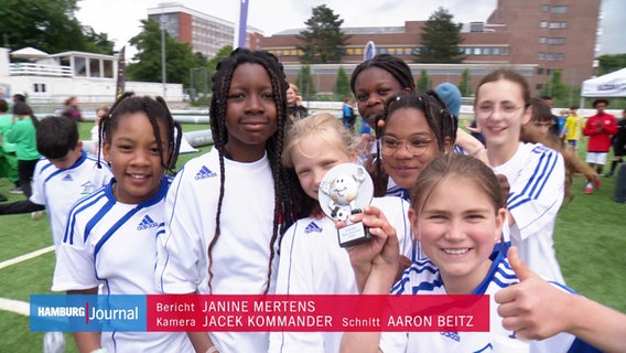 Sieben Schülerinnen in Fußball Trikots lächeln in die Kamera und präsentieren einen kleinen silbernen Pokal. © Screenshot 