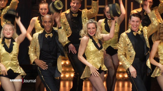 Eine große Gruppe Tänzerinnen und Tänzer auf der Bühne. Sie tragen glitzernde Outfits in schwarz und gold und lüften ihre Zilinderhüte. © Screenshot 