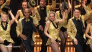 Eine große Gruppe Tänzerinnen und Tänzer auf der Bühne. Sie tragen glitzernde Outfits in schwarz und gold und lüften ihre Zilinderhüte. © Screenshot 