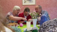 Eine Jugendliche sitzt zwischen einer Seniorin und einem Senior. Die beiden erklären ihr eine Handarbeit mit Wolle. Auf dem Tisch vor ihnen stehen Kaffee, Kuchen und Blumen. © Screenshot 