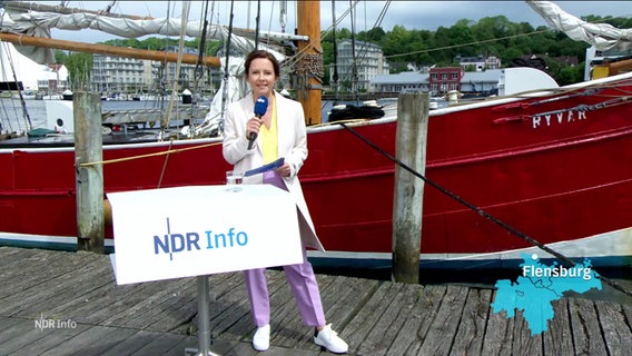 Romy Hiller moderiert draußen am Museumshafen in Flensburg. Sie steht auf einem hölzernen Kai, hinter ihr liegt ein altes Segelschiff im Wasser. © Screenshot 
