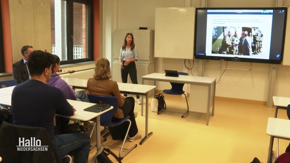 Eine 12. Klasse beim Unterricht. Vorne steht eine Lehrerin, neben ihr wird auf einem Bildschirm ein Beitrag über die Wehrdienst-Pläne angezeigt. © Screenshot 