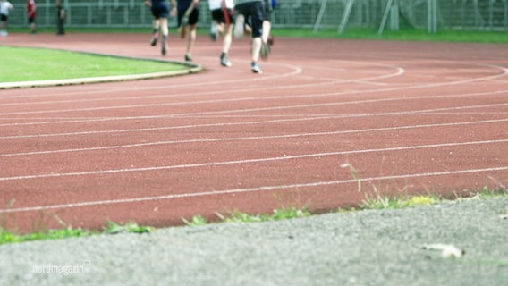 Nahaufnahme: Die Laufbahn des Stadions Lambrechtsgrund. Im Hintergrud sind die Beine einiger Läufer zu sehen. © Screenshot 