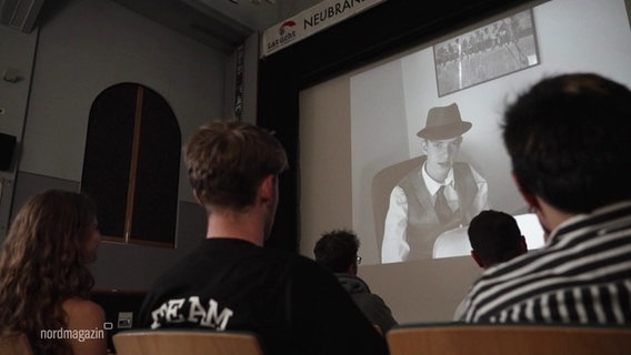 Jugendliche sitzten vor einer Kinoleinwand, auf die ein Film projeziert wird. Der Film ist in schwarz/weiß und es ist ein Jugendlicher zu sehen, der wie ein Film Noir Detektiv gekleidet ist. © Screenshot 