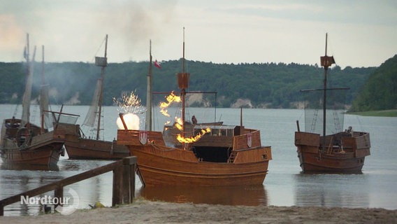 Am Ufer eines Sees treiben nachgebaute Holzschiffe, eines davon scheint zu brennen. © Screenshot 