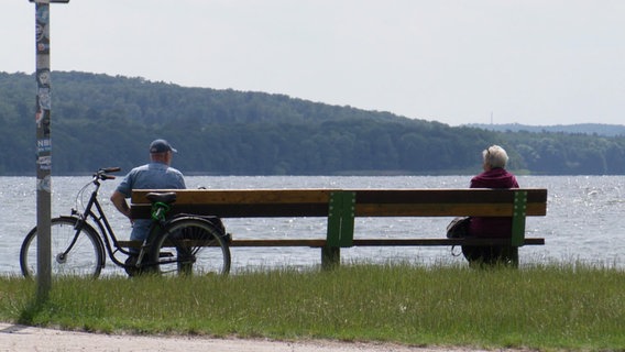 Eine Frau und ein Mann sitzen auf einer Bank, die am Ufer eines Gewässers steht. An der Bank lehnt ein Fahrrad. © Screenshot 