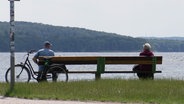 Eine Frau und ein Mann sitzen auf einer Bank, die am Ufer eines Gewässers steht. An der Bank lehnt ein Fahrrad. © Screenshot 