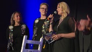 Zwei Frauen in Lederwesten mit vielen Aufnähern stehen neben einer blonden Frau mit Mikrofon auf einer Bühne. © Screenshot 
