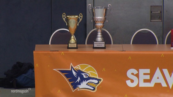 Auf einem Tisch, der mit einem Seawolves-Banner behängt ist, stehen zwei Pokale. © Screenshot 