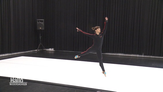 Eine Balletttänzerin in schwarzer Traininskleidung tanzt auf einem weißen Boden vor schwarzen Vorhängen. © Screenshot 